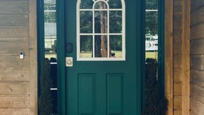 Een groene houten deur met een raam met wit kader, twee potplanten en een deurmat.