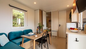A compact living space with a kitchenette, a dining area with a table and chairs, a sofa, and a window with a view of greenery. - Le Cycas, a new mobile home with sea view (Hyères)