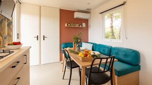 A compact living space with a kitchenette, a dining area with a table and chairs, a teal sofa, and a window with a view of greenery. - Le Cycas, a new mobile home with sea view (Hyères)