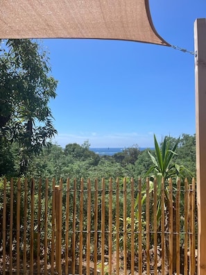 A view through a bamboo fence with a shade cloth, overlooking a lush green landscape and a distant view of the sea. - Le Cycas, a new mobile home with sea view (Hyères)