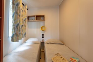 Une petite chambre à coucher, de dimensions réduites, comprenant deux lits simples, une table de chevet avec une lampe et une étagère ornée d’objets décoratifs.