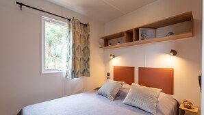 A bedroom with a bed, two bedside lamps, a window with curtains, and a shelf above the bed. - L'Olivier, mobile home with a sea view (Hyères)