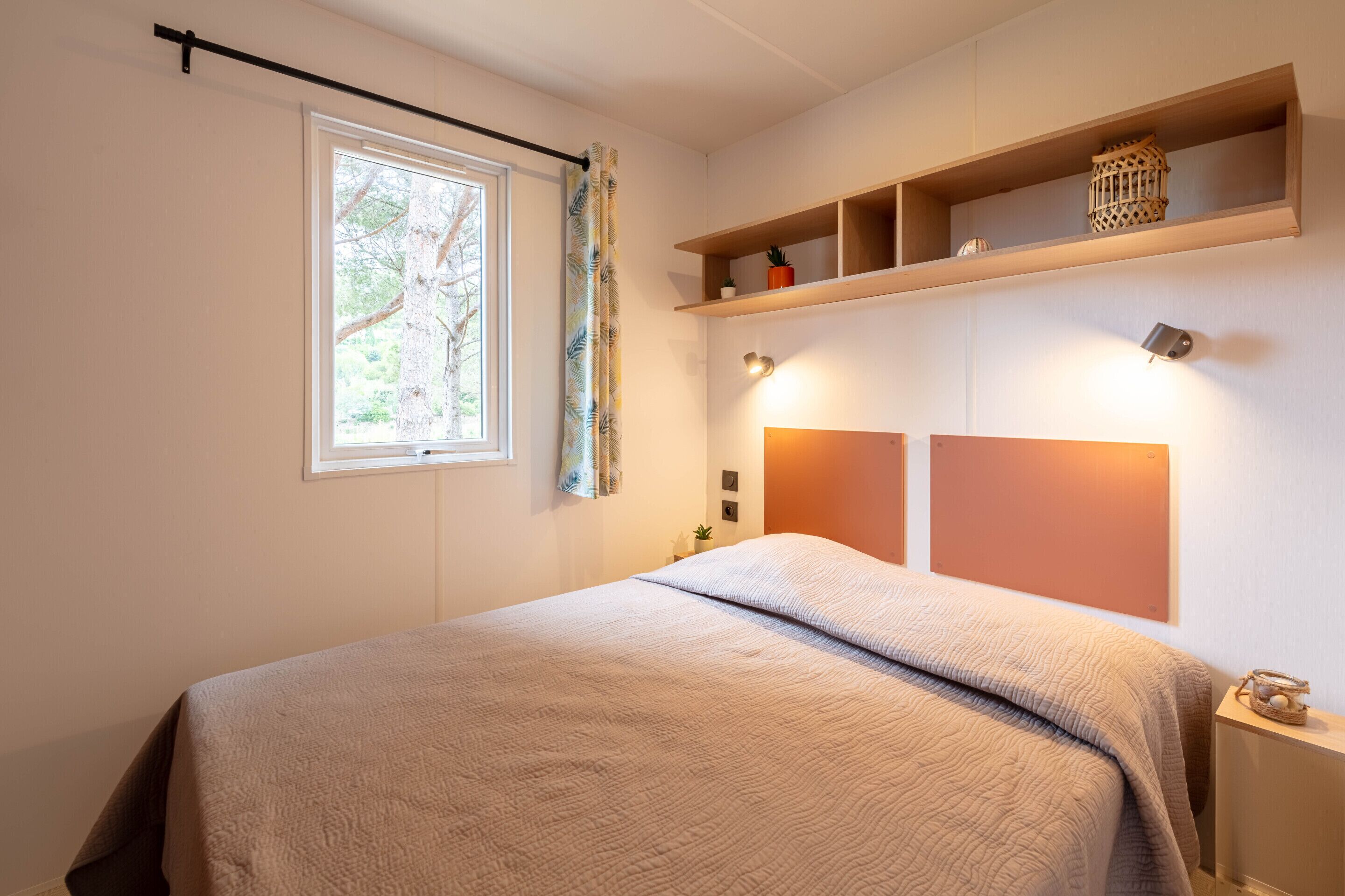 Une chambre à coucher avec un lit, une fenêtre avec des rideaux, une étagère avec des objets décoratifs et des lampes fixées au mur.