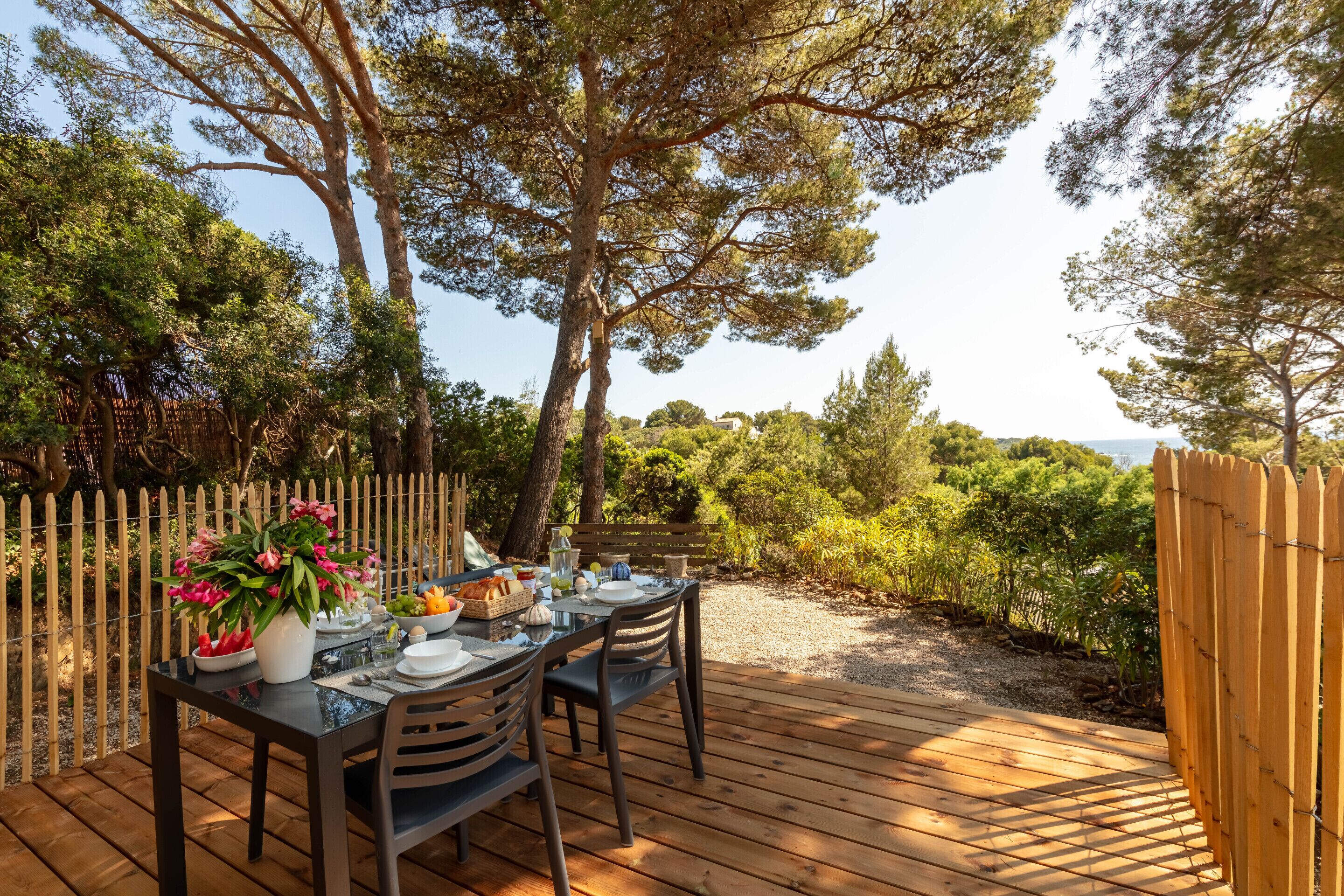 An outdoor dining area with a table set for two, surrounded by a wooden deck and a wooden fence.