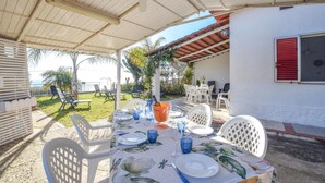 Un espace extérieur aménagé pour les repas, avec une table dressée pour quatre personnes, entourée de chaises blanches, sous une terrasse couverte offrant une vue sur la mer.