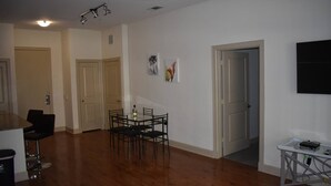 Een ruime woonkamer met houten vloer, een eettafel met stoelen, een aan de muur bevestigde televisie, en een deur die leidt naar een andere kamer.