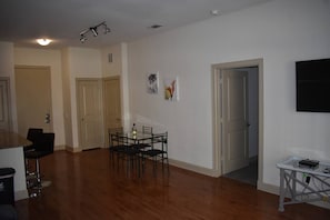 A spacious living area with a wooden floor, a dining table with chairs, a TV mounted on the wall, and a door leading to another room.