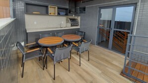 Et udendørs spiseområde med et rundt bord og stole, grill og en skydedør, der fører til en balkon.