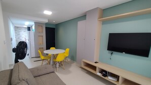 En stue med sofa, et rundt bord med gule stole, fjernsyn på væggen og en ventilator i hjørnet.