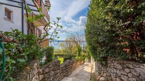Ein schmaler, gepflasterter Weg, der von üppigem Grün gesäumt wird und zu einem Gebäude mit Balkon führt, das auf ein Gewässer blickt.