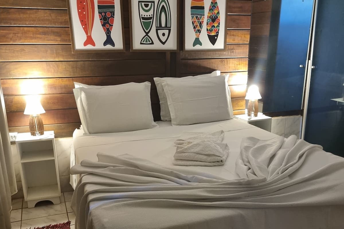 Cama bem arrumada com lençóis brancos, manta vermelha e travesseiro branco. Há duas mesas de cabeceira com abajures, uma parede de madeira com quadros e um espelho grande.