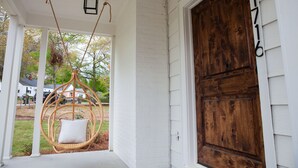 A porch with a wooden door, white siding, and a wicker swing chair with a cushion. - Beautifully Renovated and Decorated Home in Great Neighborhood (Augusta)