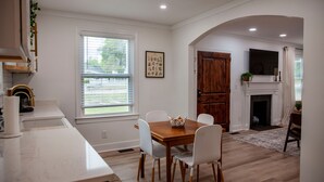 A modern kitchen with white cabinetry, a wooden dining table, and chairs. There is a window with blinds, a fireplace, and a mounted television. - Beautifully Renovated and Decorated Home in Great Neighborhood (Augusta)