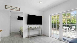 Un salon moderne avec un téléviseur à écran plat fixé au mur, une petite console sur laquelle sont disposés des objets décoratifs, un tapis à motifs et une porte-fenêtre coulissante donnant sur un espace patio extérieur.