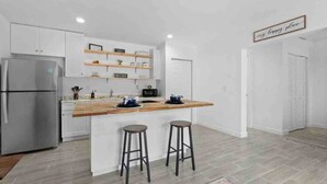 Une cuisine moderne avec des meubles blancs, un plan de travail en bois, deux tabourets et un réfrigérateur.