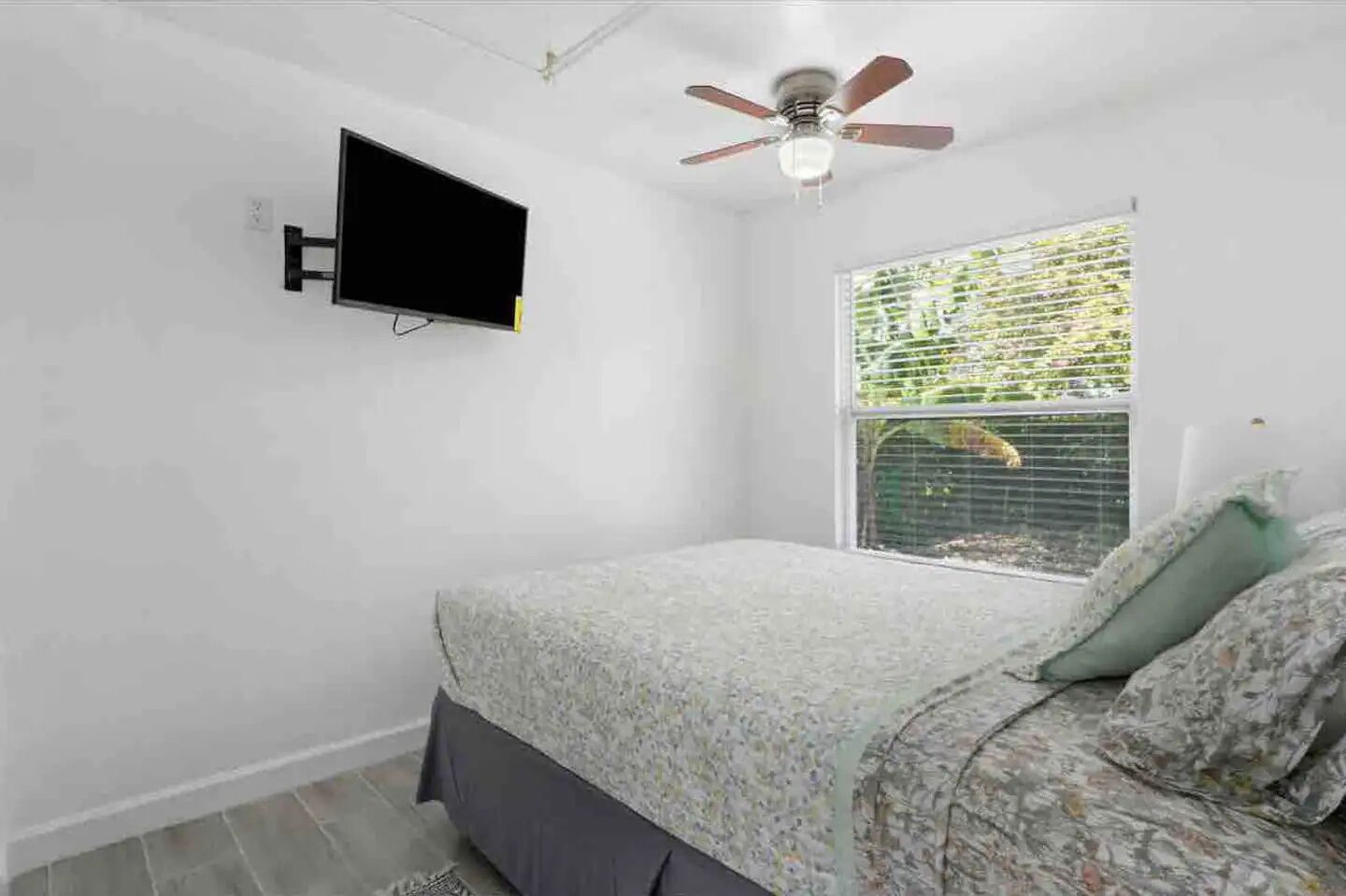 Un dormitorio con cama, ventilador de techo, televisor montado en la pared y ventana con persianas.