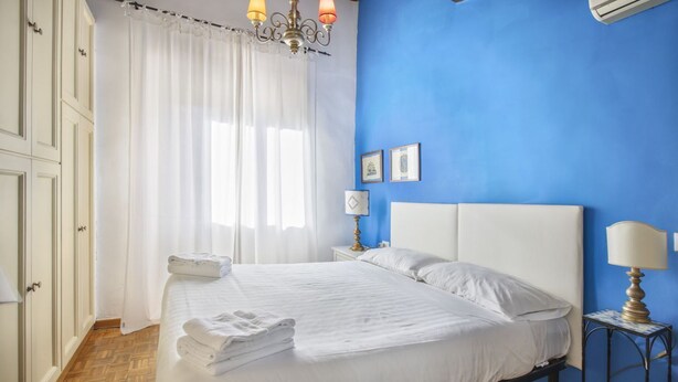 A bedroom with a bed, white curtains, a chandelier, a nightstand with a lamp, and a blue wall. - Seven House - Sweet Dreams in Florence (Firenze)