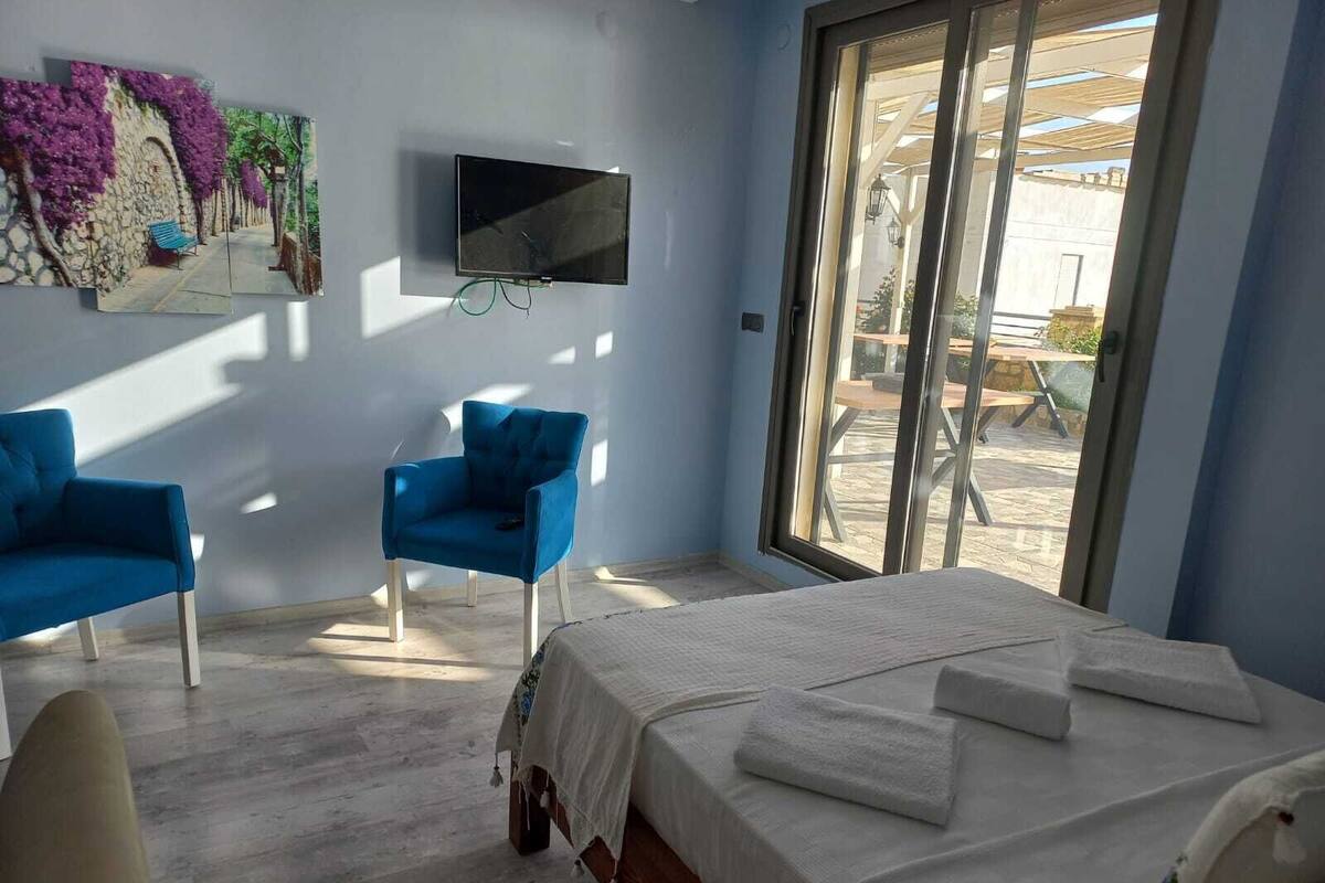 Ein Hotelzimmer mit einem Bett, zwei blauen Sesseln, einem an der Wand montierten Fernseher und einem großen Fenster mit Blick ins Freie.