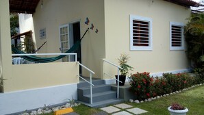 A house with a beige exterior, white window frames, a set of stairs with a white handrail, and a small garden with red flowers and a potted plant.