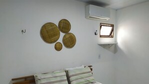 A bedroom with a bed, a wooden headboard, a wall-mounted air conditioner, a small window, and three woven wall decorations.