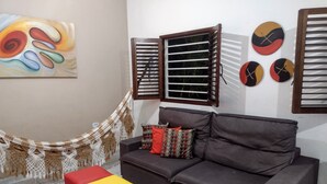 A living room with a brown sofa, a yellow ottoman, a hammock, and a colorful abstract painting on the wall.