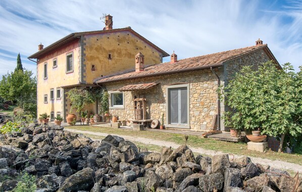 A rustic stone house with a yellow facade, a stone chimney, and a small garden with potted plants and a tree.
