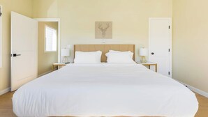 Una cama grande con ropa de cama blanca en una habitación con paredes color beige y suelo de madera.