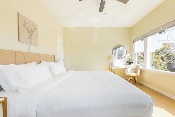 Un dormitorio de hotel con una cama grande, un ventilador de techo, una mesita de noche con lámpara, una silla y una ventana con vistas a árboles.