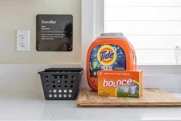 Una encimera de cocina con un recipiente de Tide PODS, una caja de suavizante Bounce y una cesta de ropa negra.
