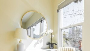 Una habitación con un espejo redondo, una silla blanca, una lámpara y un ventanal con vistas a árboles.
