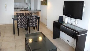 Un salon moderne avec une table basse en verre, un coin repas avec des chaises à motifs, une cuisine en arrière-plan et un téléviseur à écran plat fixé au mur.