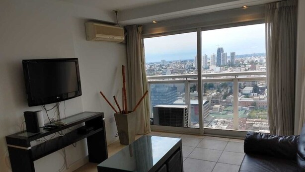 Un salon moderne avec un téléviseur à écran plat, une table basse et un canapé en cuir. Une grande fenêtre offre une vue sur la ville.