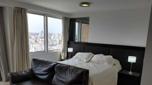 Une chambre d’hôtel moderne avec un grand lit, un canapé en cuir et une vue sur la ville par la fenêtre.