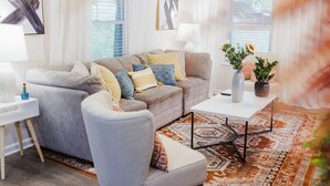 Ein Wohnzimmer mit grauem Sofa, gemusterten Kissen, einem weißen Beistelltisch, einem weißen Couchtisch und einem Teppich mit Muster.