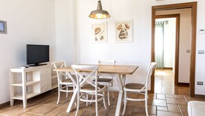 A dining area with a wooden table and white chairs, a TV on a white cabinet, and two framed pictures on the wall. - M348 - Marcelli, comfortable new three-room apartment with swimming pool and bal (Numana)