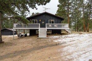 A two-story cabin with a balcony, surrounded by trees and a dirt ground.