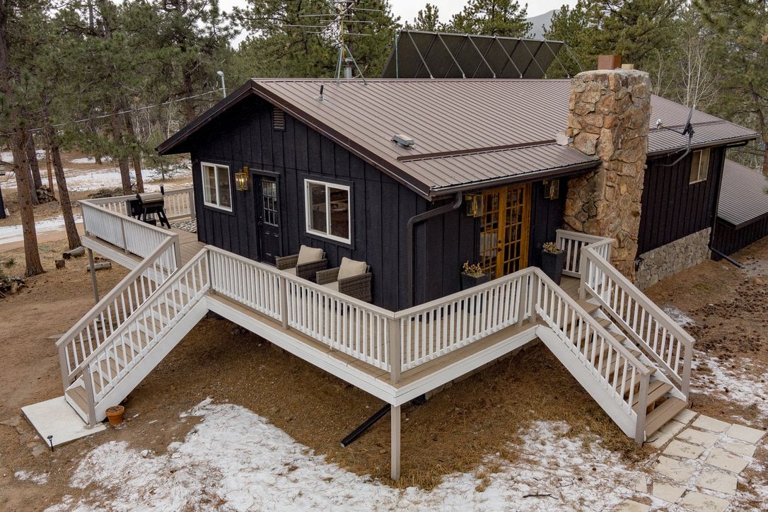 A cabin with a stone chimney, a deck with a white railing, and a small porch with outdoor seating.