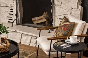 Ein gemütlicher Sitzbereich mit gepolstertem Sessel, einem kleinen runden Tisch mit Buch und Tasse sowie einem Kamin mit Holzscheiten.