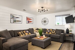 A modern living room with a sectional sofa, a coffee table, and decorative pillows. There are framed sports pennants on the wall and a mounted TV.