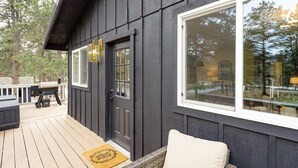 Eine dunkle Holzausfachung mit einem großen Fenster, einer Tür und einer Veranda mit Gartenmöbeln.
