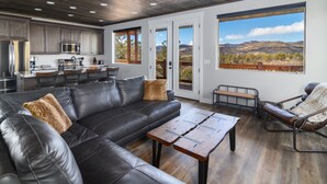 Uma sala de estar moderna com um sofá de couro, uma mesa de centro de madeira e vista para montanhas através de grandes janelas.