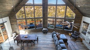 A spacious loft with a high ceiling, large windows offering views of a mountainous landscape, a dining area, and a cozy seating area with a fireplace. - Stargazer Cabin | Private Hot tub | Zion NP (Orderville)