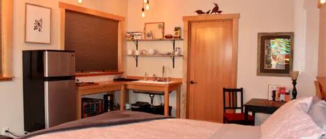 A compact room with a bed, a kitchenette, a wooden door, and a small table with a chair.