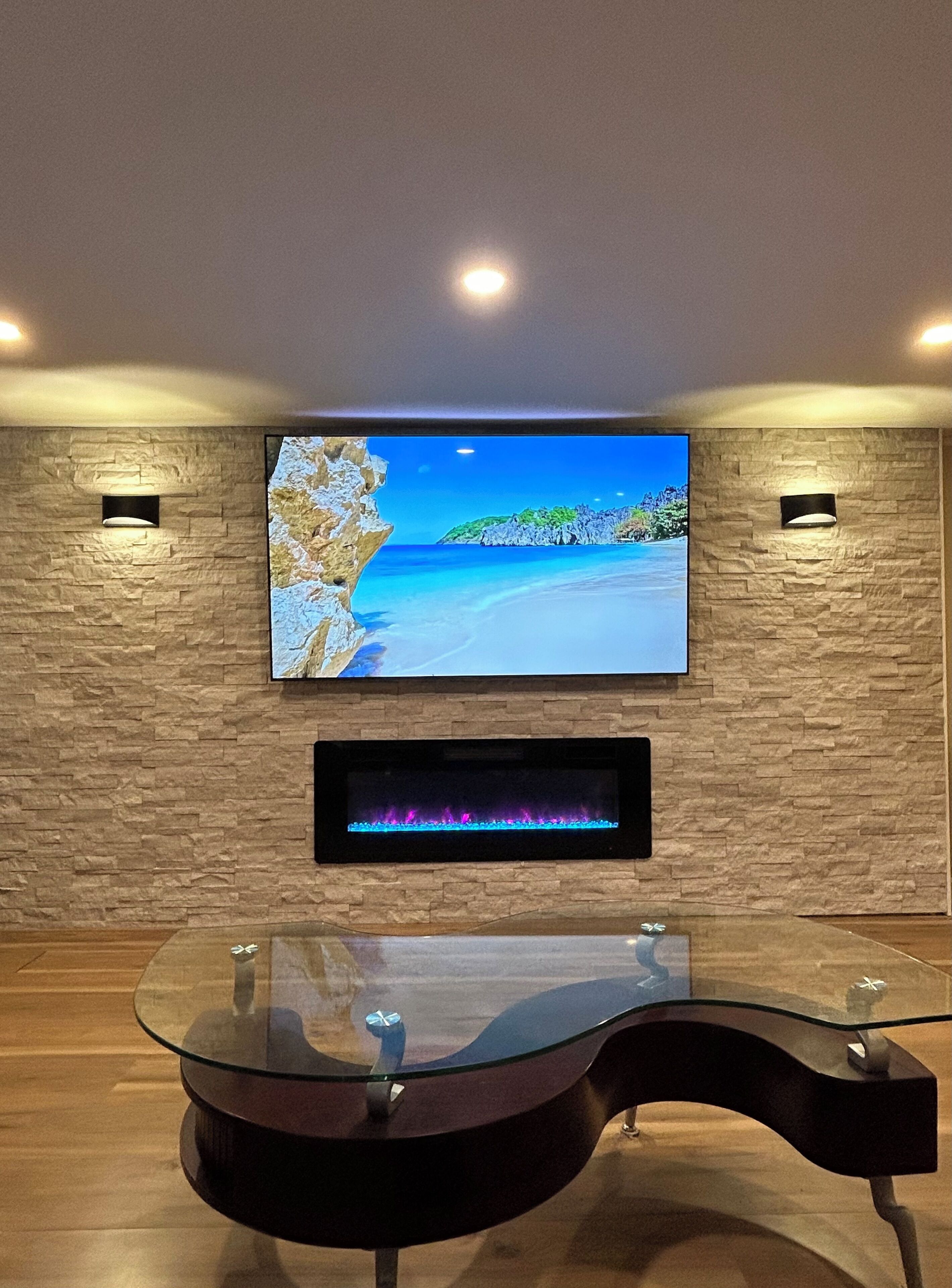 A modern living room with a flat-screen TV mounted on a stone wall, a glass-top coffee table, and a fireplace below.