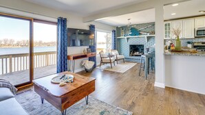 A spacious living area with a large window overlooking a body of water, a wooden coffee table, and a fireplace. - Spacious Lakefront Home in White Lake (White Lake Charter Township)