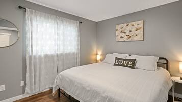 Een slaapkamer met een bed, nachtkastje, een wandlamp, een raam met gordijnen en een kunstwerk aan de muur.