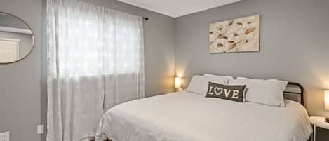 A bedroom with a bed, a nightstand, a wall-mounted lamp, a window with curtains, and a wall art piece.