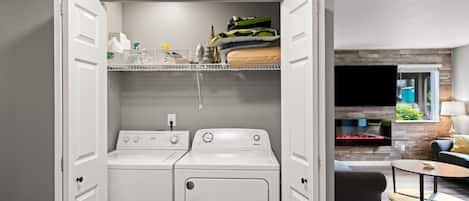 A laundry room with a washer and dryer, a shelf with folded clothes, and a view into a living area with a fireplace.