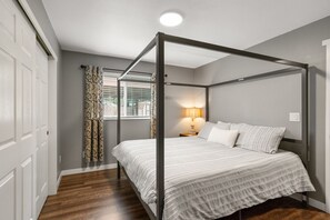 Una cama con dosel con una colcha blanca, una lámpara de noche y una ventana con cortinas estampadas.