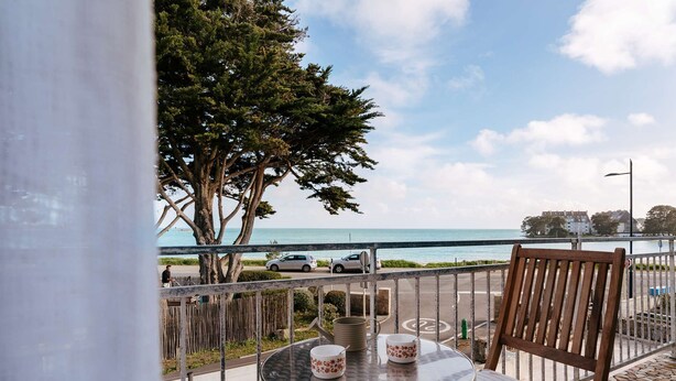 Een balkon met een tafel en stoel, met uitzicht op een strand en een weg met geparkeerde auto's.